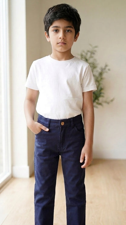A young boy standing indoors wearing a plain white short-sleeve crewneck t-shirt and dark navy blue pants, with his hands in his pockets.